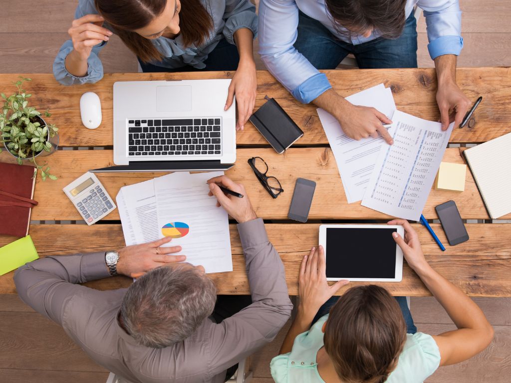 Vista aérea de cuatro profesionales colaborando en una mesa de madera con documentos, gráficos y un computador, representando la gestión de tareas y la optimización de recursos, se encuentran buscando sobre qué es el outsourcing.