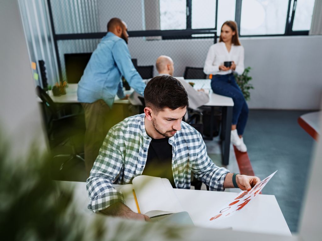 Trabajador de recursos humanos concentrado revisando gráficos y documentos en su escritorio mientras otros compañeros trabajan en una oficina. La escena representa el outsourcing de remuneraciones